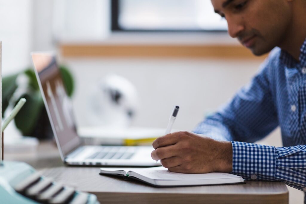 Man writing on notebook researching types of keywords. A laptop is next to the notebook.