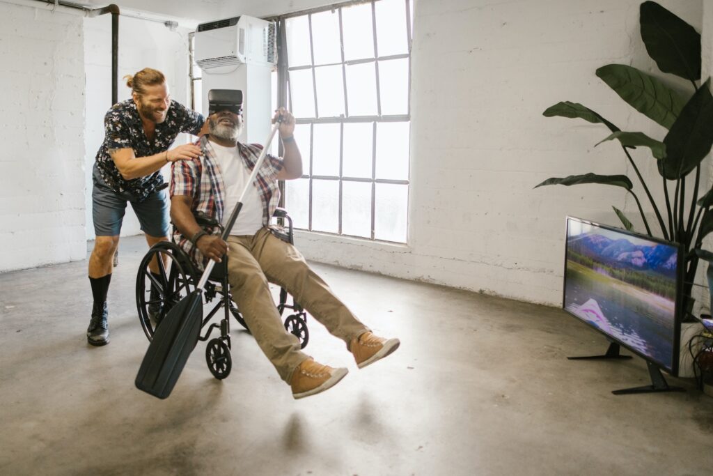 A man pushing another man in a wheelchair with a virtual reality headset on. Man in wheelchair holding a paddle.