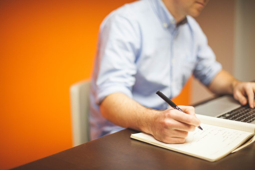 A man sits at a desk. He is typing something on a laptop and simultaneously writing in a notebook.